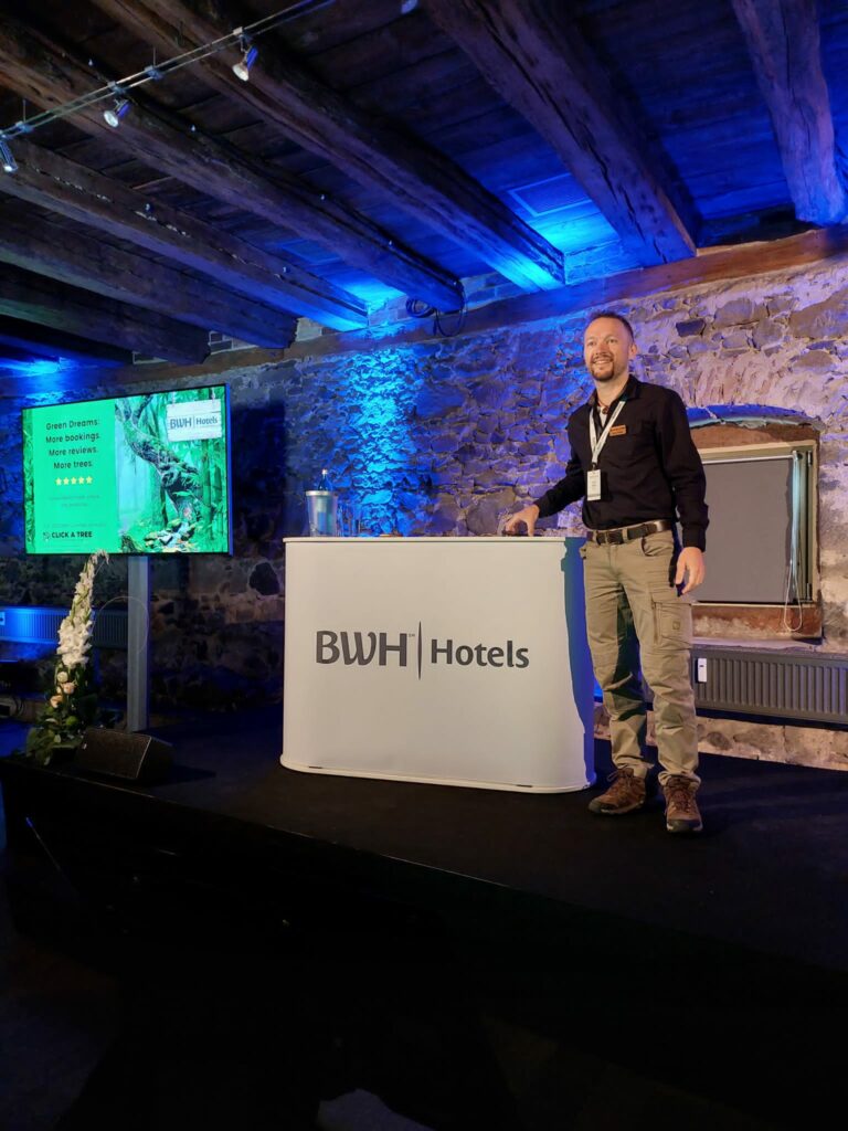 Chris Kaiser steht vor einem Pult, dem Publikum zugewandt, und berichtet von Nachhaltigkeit im Hotel. Der raum ist rustikal mit unverputzten Steinwänden und atmosphärischem blauen licht.